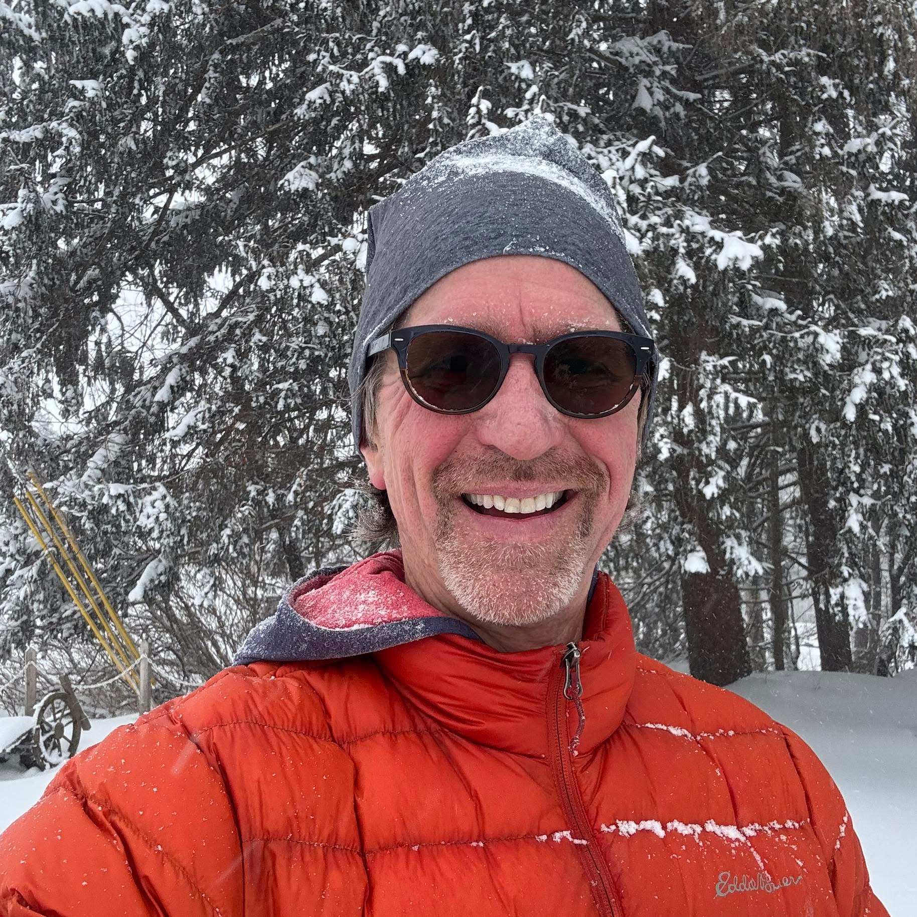 David smiling in a snowy forest, wearing an orange jacket and beanie